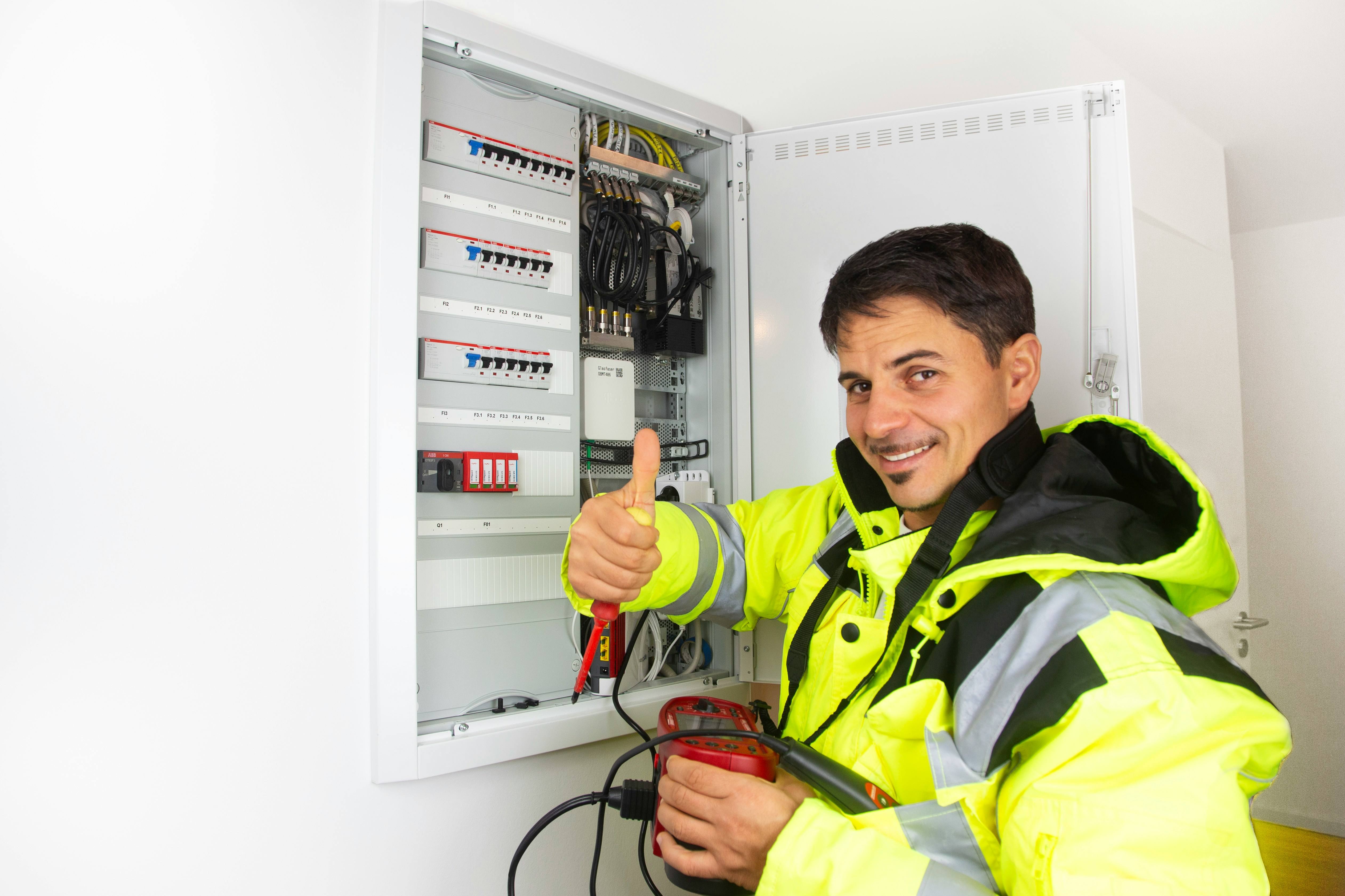 Iemand in een felgele werkjas bij een geopende groepenkast, met een meetapparaat in de hand.